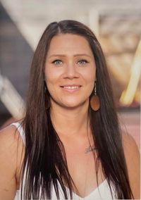 Eine Frau mit langen dunklen Haaren und blauen Augen lächelt in die Kamera. Sie trägt ein weißes ärmelloses Oberteil und hat eine Tätowierung auf ihrer Brust.