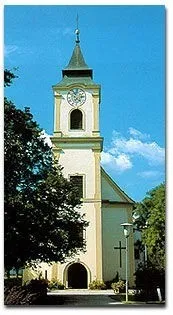 Eine Kirche mit einem hohen Turm und einer Uhr zeigt sich vor einem blauen Himmel mit verstreuten Wolken. Bäume und ein Kreuz sind vor der Kirche sichtbar.