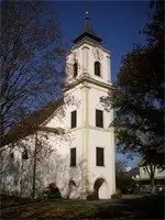 Eine weiße Kirche mit einem braunen Dach und einem hohen Turm, umgeben von Bäumen unter einem klaren blauen Himmel.