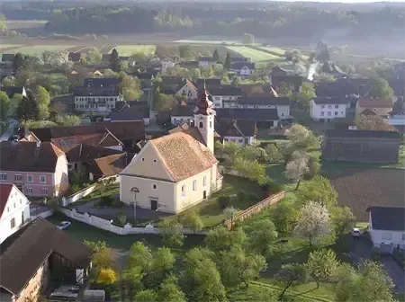 Eine Luftaufnahme eines Dorfes mit vielen Häusern, einer Kirche und Bäumen. Ein Auto ist vor der Kirche geparkt.