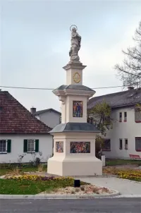Ein Denkmal mit einer Statue einer Person an der Spitze ist von Pflanzen und Blumen umgeben. Im Hintergrund befinden sich Häuser mit Fenstern und Schornsteinen.