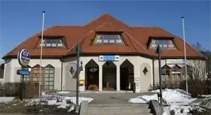Ein großes Gebäude mit rotem Dach und mehreren Fenstern, es hat ein Schild mit blauem Text und einem Bogeneingang. Es gibt Stufen, die zum Eingang führen, mit Schnee an den Seiten.