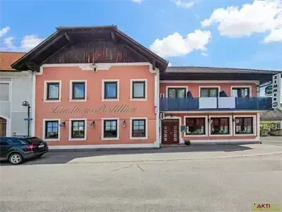 Ein zweistöckiges Gebäude mit braunem Dach und rosa Wänden, benannt Landhaus Postillion, hat einen Balkon und mehrere Fenster. Ein schwarzes Auto ist davor geparkt.