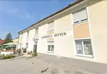 Ein Gebäude mit 'GASTRONOMIE RITTER' darauf geschrieben, es hat einen Parkplatz davor mit Pflanzen und einem Schirm an der Seite.
