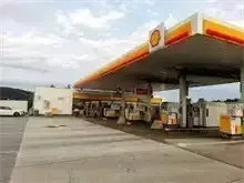 Eine Shell-Tankstelle mit gelben und roten Überdachungen, mehreren Zapfsäulen und einem geparkten weißen Auto in der Nähe.