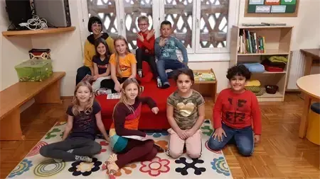 Eine Gruppe von Kindern in einem Raum, einige sitzen auf einer roten Couch, andere auf dem Boden, mit einem Holzschreibtisch und Regalen im Hintergrund.