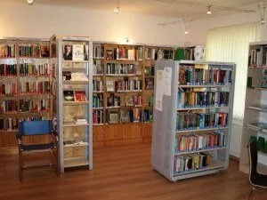 Ein Innenansicht einer Bibliothek mit Holzboden, mehreren Bücherregalen voller Bücher und zwei blauen Stühlen. Die Regale haben Glasfronten und Holzrücken. Ein weißer Heizkörper ist an der Wand sichtbar.