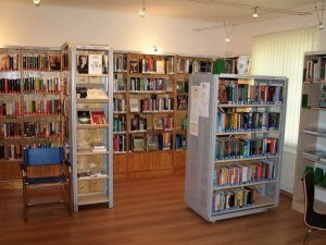 Ein Innenansicht einer Bibliothek mit Holzboden, mehreren Bücherregalen voller Bücher und zwei blauen Stühlen. Die Regale haben Glasfronten und Holzrücken. Ein weißer Heizkörper ist an der Wand sichtbar.