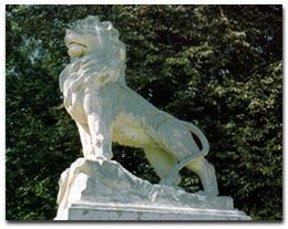 Eine weiße Löwenstatue steht auf einem Felsen vor einer Kulisse aus üppigem grünem Laub. Der Löwe scheint bereit zu sein, möglicherweise brüllend, mit einem ruhigen Ausdruck.