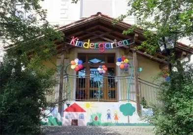 Der Eingang zu einem Kindergartengebäude zeigt eine farbenfrohe Wandmalerei, Luftballons und ein Schild mit der Aufschrift 'Kindergarten'.