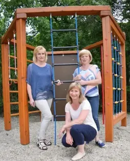Drei Frauen posieren für ein Foto im Spielplatz, eine steht auf einer Leiter und die anderen knien. Die Spielstruktur hat bunte Stangen und eine rote Tür.
