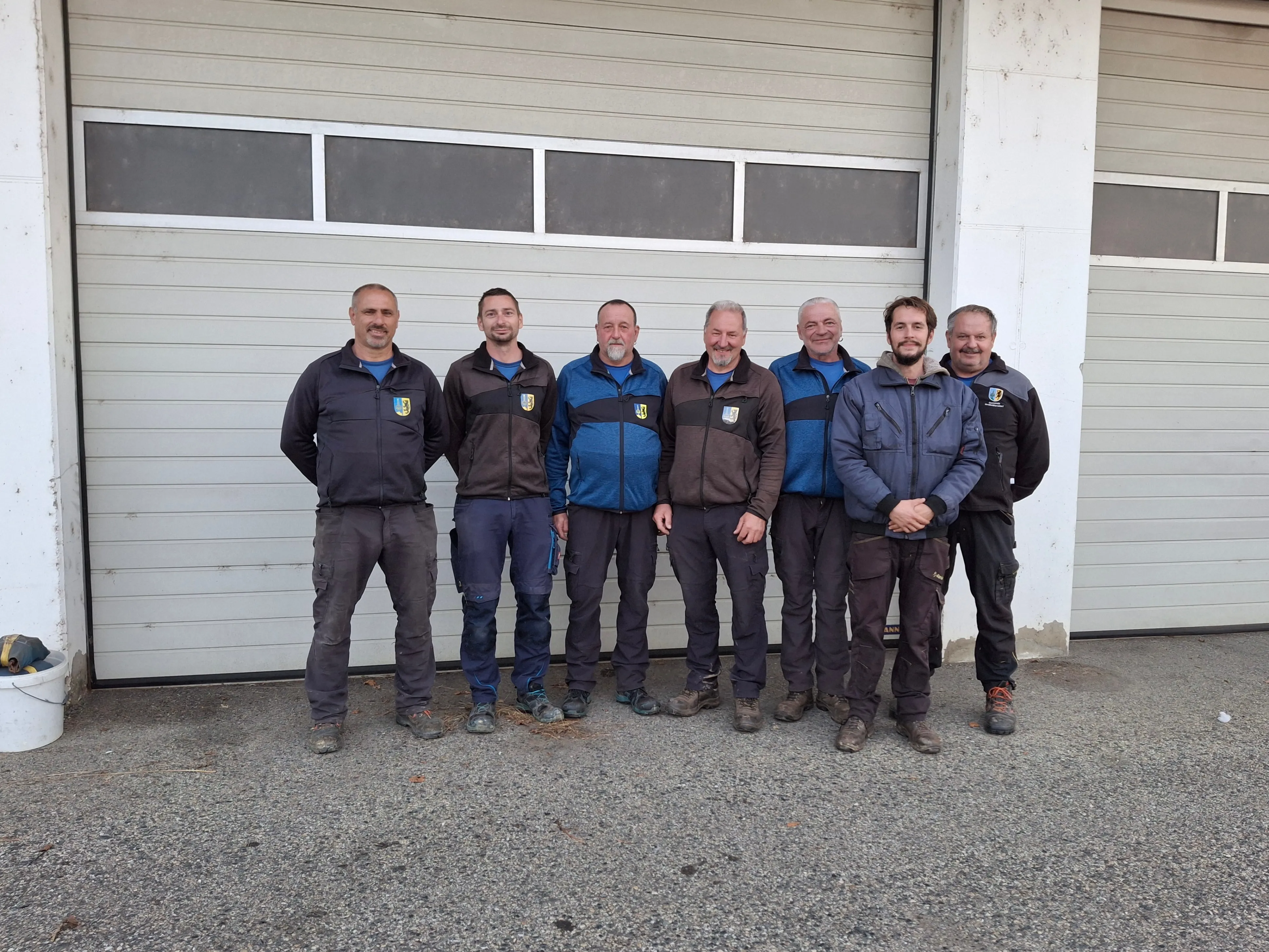 Sechs Männer stehen zusammen vor einem Gebäude. Sie tragen Jacken und Hosen, die für die Arbeit im Freien geeignet sind. Im Hintergrund befindet sich eine Garage mit Fenstern.