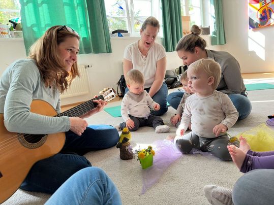 Eine Frau spielt Gitarre, während eine Gruppe von Müttern und ihren Babys auf dem Boden sitzen, einige halten Blumen.