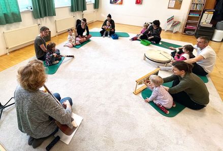 Eine Gruppe von Erwachsenen und Kindern sitzt im Kreis auf dem Boden und spielt Musikinstrumente. Die Erwachsenen spielen Gitarren und Trommeln, während die Kinder Xylophon spielen.