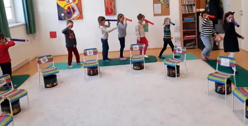 Eine Gruppe von Kindern steht in einem Raum, jeder hält eine rote Röhre, und ein Erwachsener hält ebenfalls eine rote Röhre. Die Kinder tragen Turnschuhe und der Raum hat einen weißen Teppich und bunte Stühle.