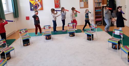 Eine Gruppe von Kindern steht in einem Raum, jeder hält eine rote Röhre, und ein Erwachsener hält ebenfalls eine rote Röhre. Die Kinder tragen Turnschuhe und der Raum hat einen weißen Teppich und bunte Stühle.
