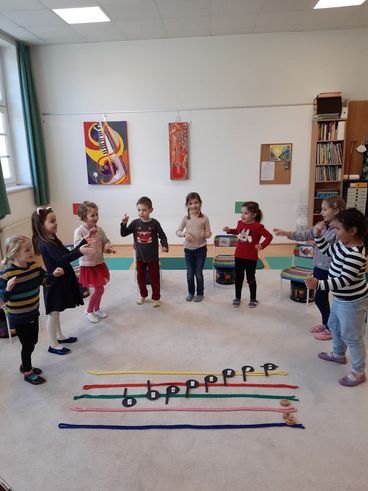 Eine Gruppe junger Kinder steht in einem Kreis, wahrscheinlich in einem Klassenzimmer, und spielt mit Musikinstrumenten. Eine bunte Musiknotenlinie liegt auf dem Boden.