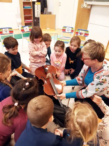 Eine Frau mit einem Blumenpullover unterrichtet eine Gruppe von Kindern über ein Cello in einem Klassenzimmer.