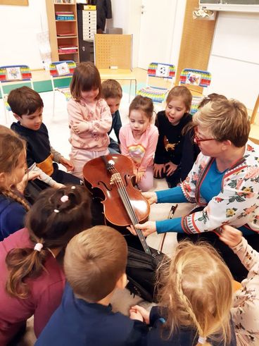 Eine Frau mit einem Blumenpullover unterrichtet eine Gruppe von Kindern über ein Cello in einem Klassenzimmer.
