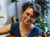 Eine Frau mit Brille und einer Halskette lächelt, während sie eine Gitarre spielt. Hinter ihr hängen zwei Gitarren an der Wand und eine Topfpflanze.