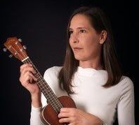 Eine Frau mit langen Haaren hält ein kleines Ukulele in den Händen und schaut konzentriert auf das Instrument. Sie trägt ein weißes Langarmshirt.