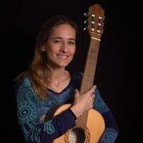 Eine Frau mit langen Haaren lächelt und hält eine klassische Gitarre in beiden Händen. Sie trägt ein blaues Langarmshirt.