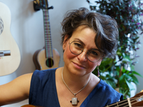 Eine Frau mit kurzen Haaren, die eine Brille und eine Halskette trägt, lächelt und spielt Gitarre. Hinter ihr hängen drei Gitarren an der Wand.