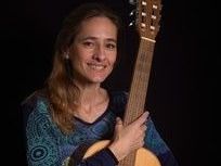 Eine Frau mit langen Haaren hält eine Gitarre und lächelt in die Kamera, trägt ein blaues gemustertes Oberteil.