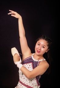 Eine Frau in einem bunten Kleid und weißen Handschuhen posiert mit einem Ballettschuh in der Hand vor schwarzem Hintergrund.