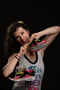 Eine Frau mit lockigem Haar lächelt und hält ein Paar bunte Turnschuhe in der Hand, sie trägt ein weißes Tanktop mit dem Text 'LIFE is better WHEN YOU'.