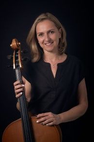 Eine Frau mit blondem Haar lächelt und hält ein Cello in beiden Händen. Sie trägt ein schwarzes Oberteil und eine Halskette. Das Cello hat einen braunen Korpus und einen schwarzen Hals. Der Hintergrund ist schwarz.
