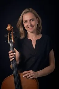Eine Frau mit blondem Haar lächelt und hält ein Cello in beiden Händen. Sie trägt ein schwarzes Oberteil und eine Halskette. Das Cello hat einen braunen Korpus und einen schwarzen Hals. Der Hintergrund ist schwarz.