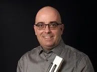Ein kahlköpfiger Mann mit Brille lächelt und hält ein Musikinstrument in der linken Hand. Er trägt ein graues Hemd.