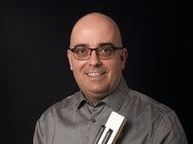 Ein kahlköpfiger Mann mit Brille lächelt und hält ein Musikinstrument in der linken Hand. Er trägt ein graues Hemd.