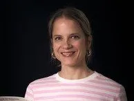 Eine Frau mit kurzen Haaren, die ein rosa-weiß gestreiftes Shirt trägt, lächelt fröhlich in die Kamera.