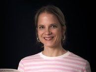 Eine Frau mit kurzen Haaren, die ein rosa-weiß gestreiftes Shirt trägt, lächelt fröhlich in die Kamera.