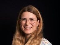 Eine Frau mit langen blonden Haaren, die eine Brille trägt und hell lächelt vor einem schlichten schwarzen Hintergrund.