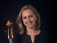 Eine Frau mit kurzen blonden Haaren lächelt, während sie ein Cello hält, eine schwarze Bluse und eine Halskette trägt.