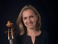 Eine Frau mit kurzen blonden Haaren lächelt, während sie ein Cello hält, eine schwarze Bluse und eine Halskette trägt.