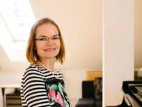 Eine Frau mit kurzen Haaren und Brille steht vor einem Klavier und lächelt für ein Foto. Sie trägt ein schwarz-weiß gestreiftes Shirt mit Blumenmuster. Hinter ihr sind ein Schreibtisch und ein schwarzer Stuhl sowie ein Oberlicht zu sehen.