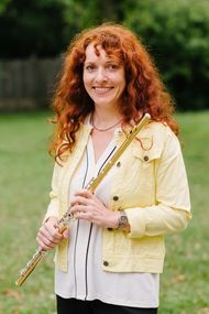 Eine Frau mit lockigem rotem Haar lächelt und hält eine Flöte in einem Park. Sie trägt eine gelbe Jacke und ein weißes Hemd.