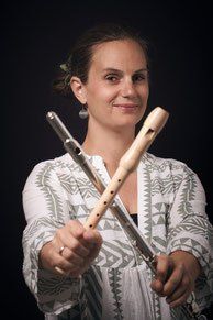 Eine Frau lächelt und hält zwei Holzflöten in den Händen. Sie trägt ein weißes Oberteil mit geometrischem Muster.