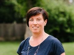 Eine Frau mit kurzen Haaren lächelt und steht in einem Grasbereich mit einem Holzzaun und Bäumen im Hintergrund. Sie trägt ein blaues Shirt und eine silberne Halskette.