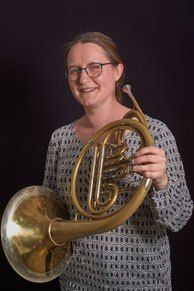 Eine Frau mit Brille und gemustertem Oberteil hält ein großes Messing-Waldhorn in beiden Händen und lächelt in die Kamera.