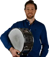 Ein Mann mit Bart hält eine Snare-Trommel in den Händen. Er trägt ein blaues Hemd.