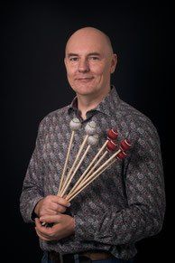 Ein kahlköpfiger Mann mit einem Lächeln hält eine Reihe von Schlägeln, wahrscheinlich für Percussion, vor einem dunklen Hintergrund.