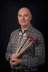 Ein kahlköpfiger Mann mit einem Lächeln hält eine Reihe von Schlägeln, wahrscheinlich für Percussion, vor einem dunklen Hintergrund.