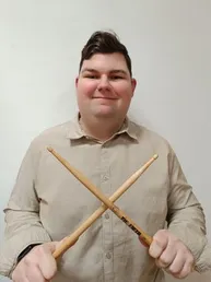 Ein Mann mit fröhlichem Ausdruck hält zwei Drumsticks in beiden Händen, gekleidet in einem beigen Hemd.