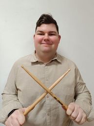 Ein Mann mit fröhlichem Ausdruck hält zwei Drumsticks in beiden Händen, gekleidet in einem beigen Hemd.