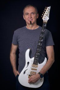 Ein Mann mit einer Zigarette im Mund hält eine weiße E-Gitarre mit komplizierten Designs. Er trägt ein graues T-Shirt und eine schwarze Armbanduhr.